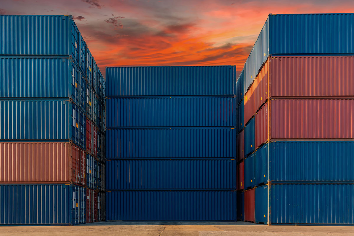 Tall stacks of blue and red shipping containers in a container yard under an orange and red sunset sky