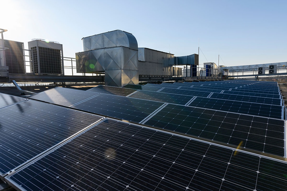 Rows of solar panels on a flat commercial roof beside large metal ventilation ducts and rooftop air conditioning units