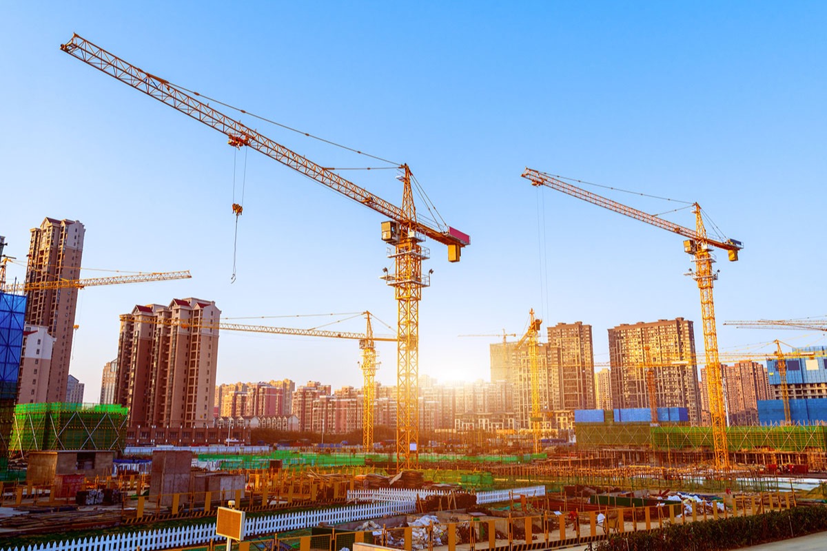 Yellow tower cranes over a large construction site with high-rise apartment blocks behind, under a clear sky with a low, bright sun