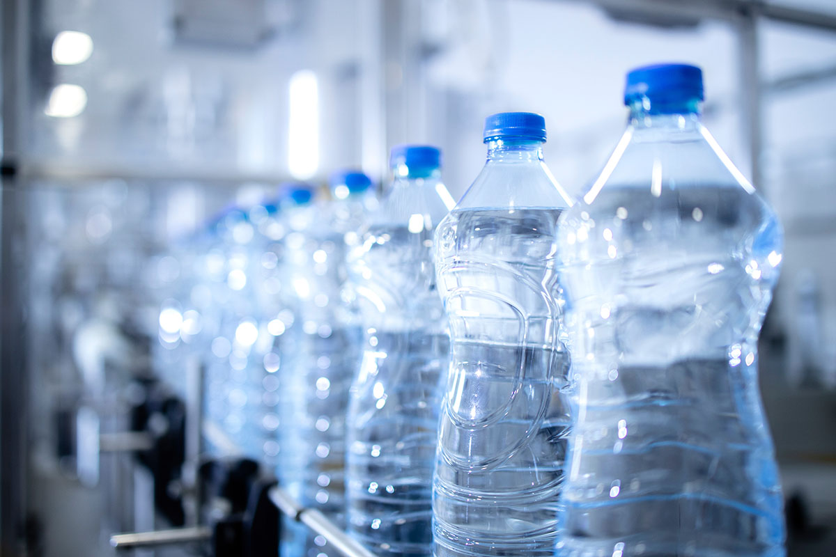A row of clear plastic water bottles with blue caps moving along a conveyor on a bottling line, the nearest bottles in sharp focus