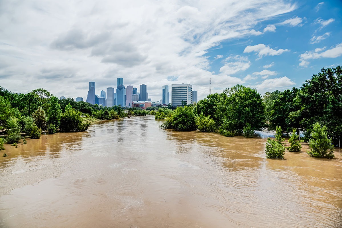 A swollen, muddy bayou running high through flooded trees toward the downtown Houston skyline under a partly cloudy sky