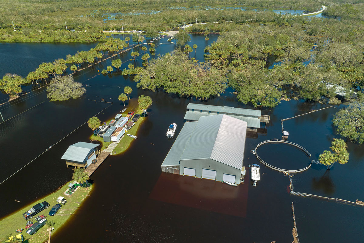 Aerial view of a metal warehouse and outbuildings standing in brown floodwater after a hurricane, with boats moored beside the buildings and flooded woodland stretching to the horizon