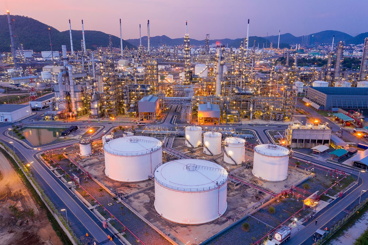 Aerial view of an oil refinery at dusk, with large white storage tanks in the foreground and lit processing towers in front of forested hills