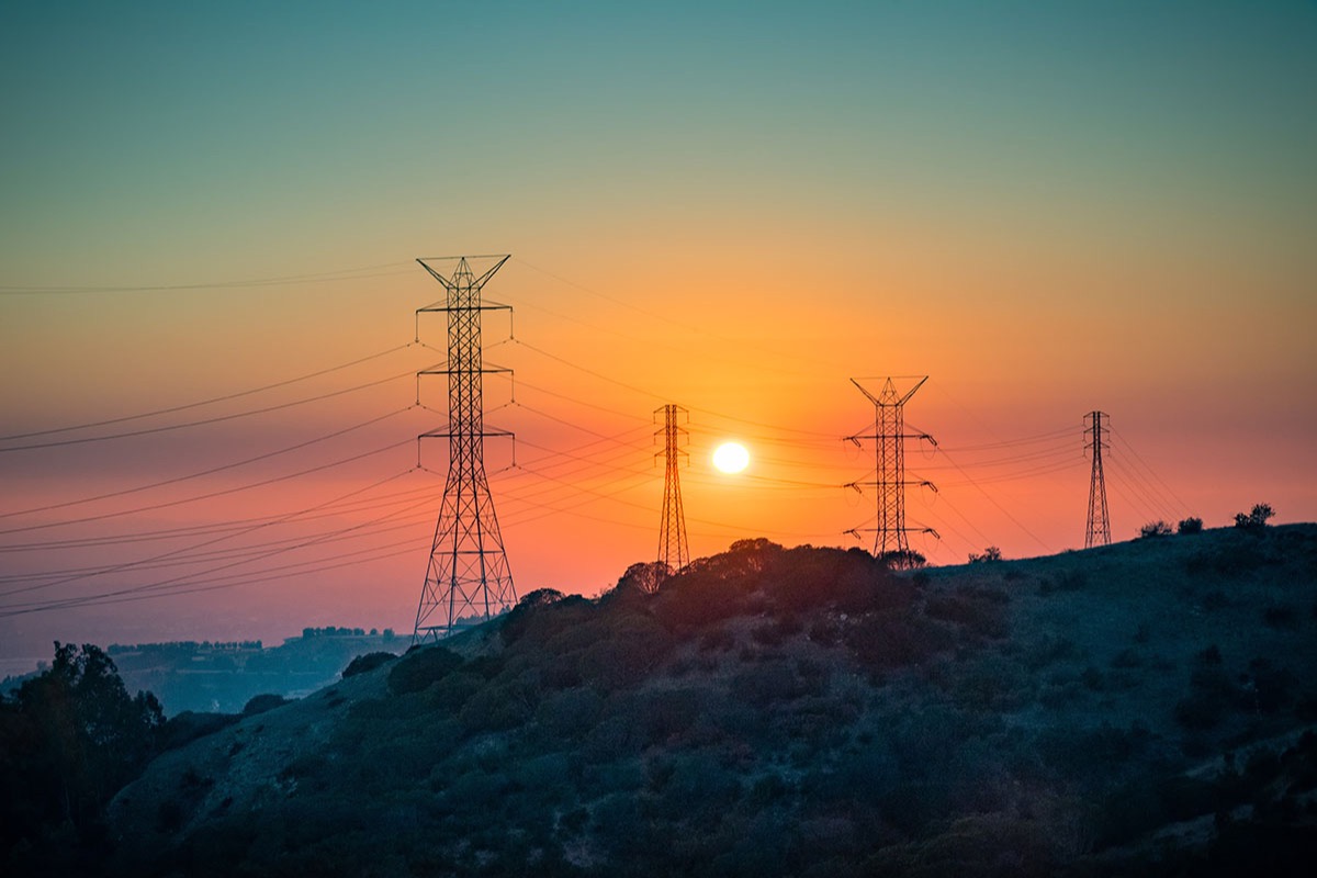 Silhouetted high-voltage transmission towers and power lines on a scrub-covered hillside in the San Gabriel Mountains against an orange sunset sky